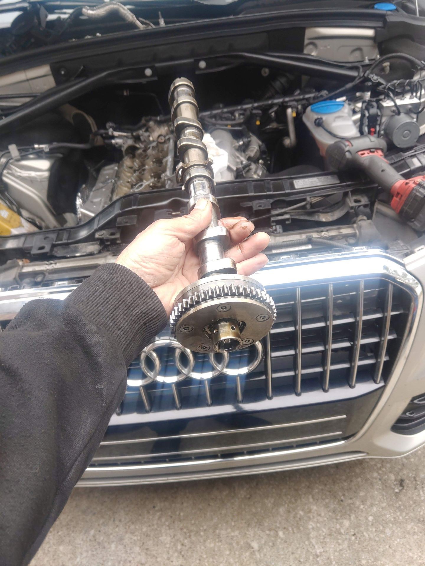 Person holding a camshaft in front of an open car hood with an engine visible inside.