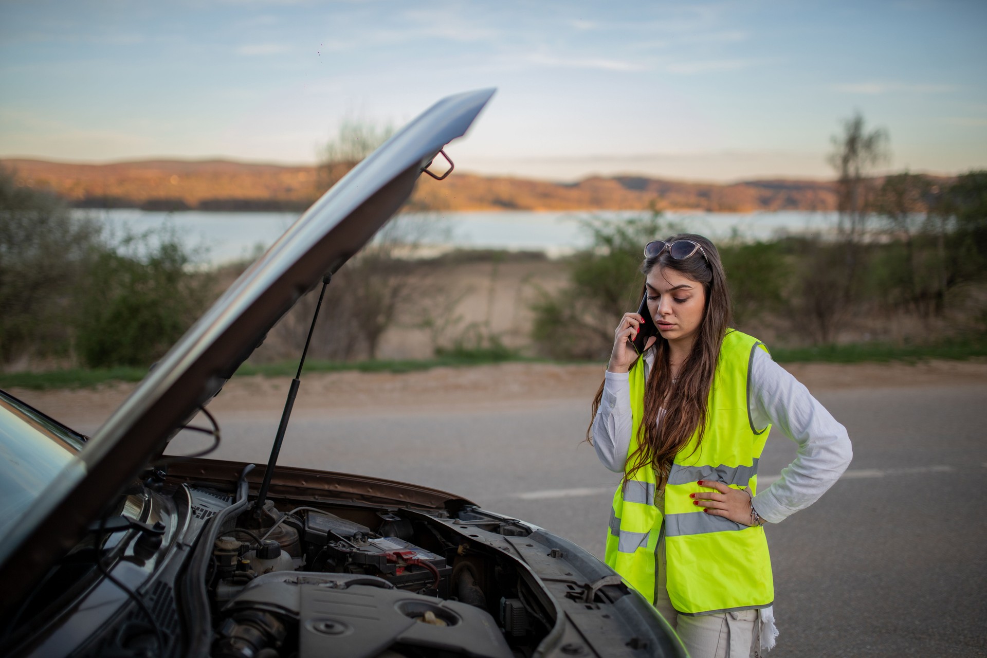 Young stressed woman standing next to her broke down car and using her phone to call for help